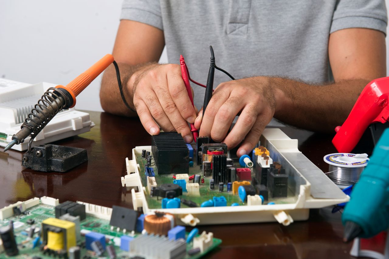 services-03 Man working on an electronic circuit board with tools on a table, showing close-up of hands and components.